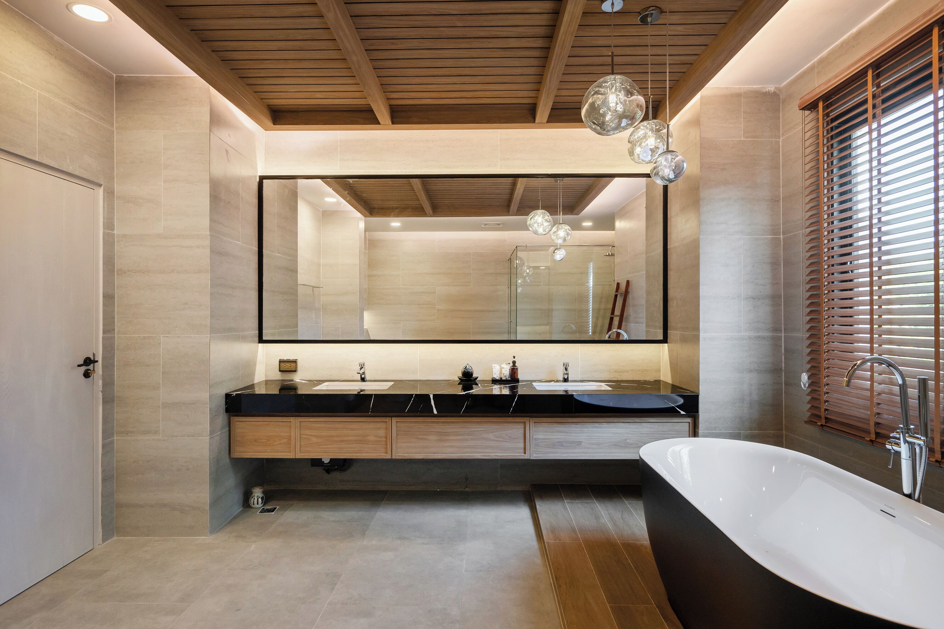 A modern bathroom with a large mirror, two sinks, a freestanding bathtub, wooden ceiling beams, and a large window with blinds.