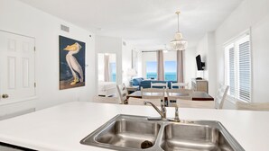 A modern kitchen with a double sink, white cabinetry, and a view of the living area with a dining table and chairs.