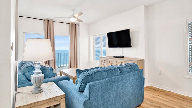 A living room with a sofa, a table with a lamp, a ceiling fan, a TV, and a window with blinds.