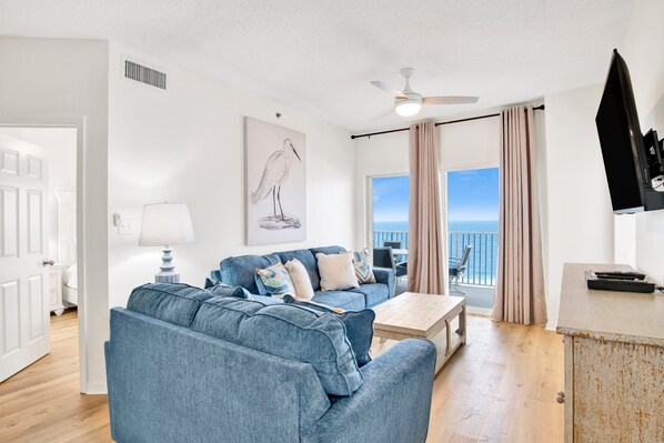 A living room with a blue sofa, a coffee table, and a painting of a bird. There is a ceiling fan, a TV, and a view of the ocean through large windows.