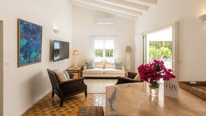 A spacious living room with a sofa, a coffee table, and a TV. There is a window with a view of greenery outside.