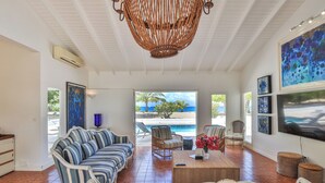 Amplia sala de estar con vista a la playa y la piscina, con sofás a rayas, sillas de mimbre y un televisor grande.