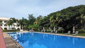 Una piscina con sillas de descanso y sombrillas, rodeada de vegetación y un edificio al fondo.