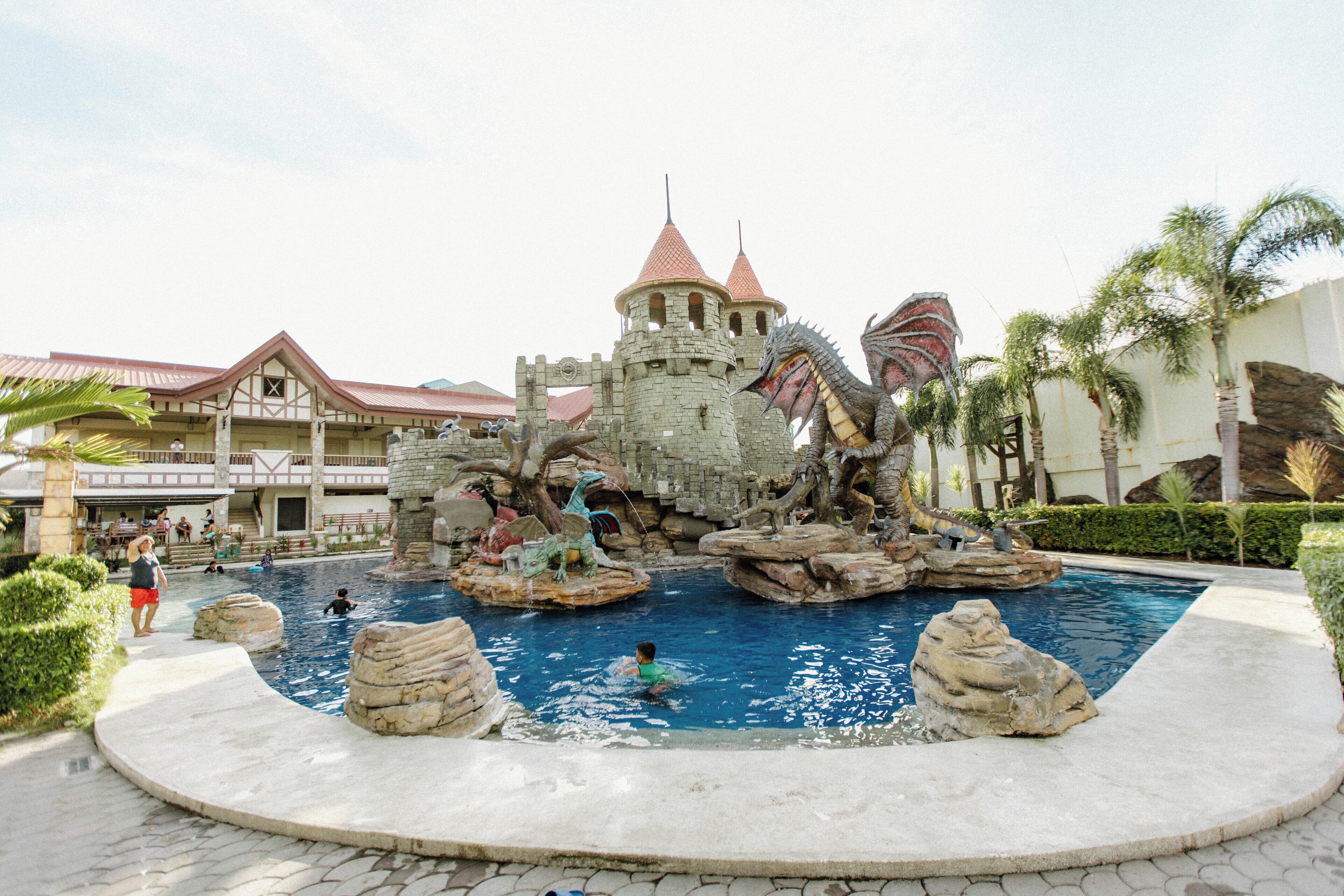 A castle-themed water feature with a pool, surrounded by palm trees and a building with a red roof in the background.