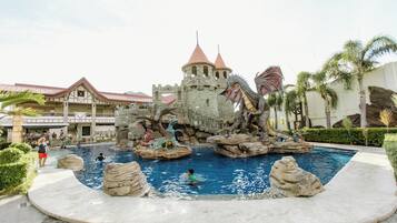 A castle-themed water feature with a pool, surrounded by palm trees and a building with a red roof in the background.