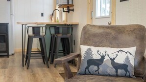 A cozy living room with a brown armchair and a cushion featuring deer silhouettes. A wooden dining table with chairs is in the background. - Hill Side Suite (Little Valley)