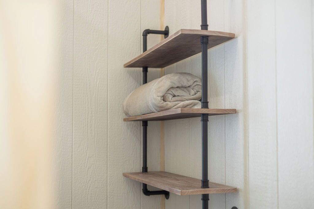 A corner shelf unit with three wooden shelves and a rolled-up blanket on the middle shelf.