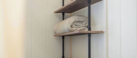 A corner shelf unit with three wooden shelves and a rolled-up blanket on the middle shelf.