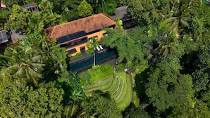 An aerial view of a tropical resort with a swimming pool, lush greenery, and a large house with a tiled roof.