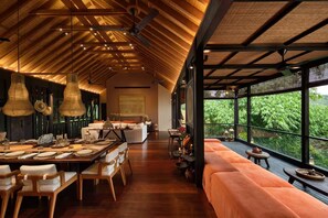 A spacious dining area with a long table set for a meal, a cozy seating area with cushions, and large windows offering a view of lush greenery. - Sumeru Botanical Estate (Ubud)