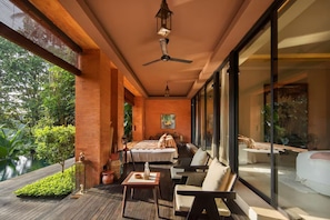 A spacious room with a bed, a seating area with a table, and large windows offering a view of the outdoors. - Sumeru Botanical Estate (Ubud)