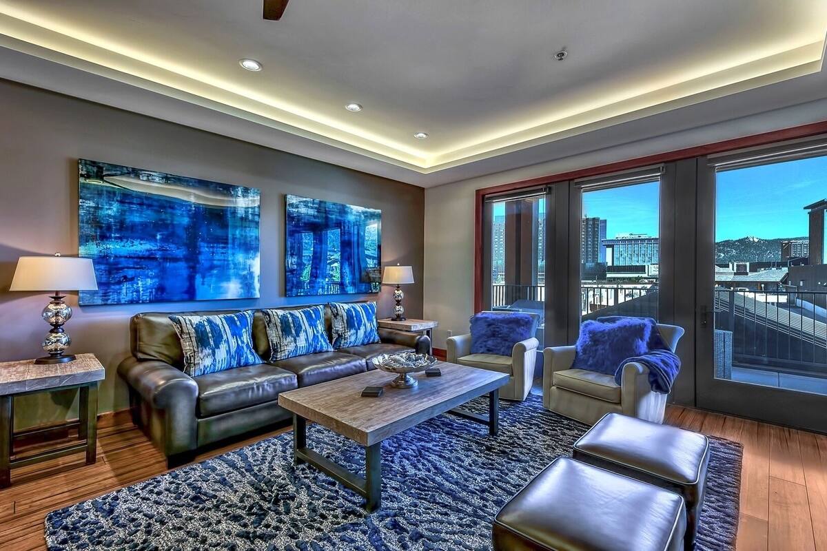 A modern living room with a leather sofa, a wooden coffee table, and a blue patterned rug. There are two floor lamps and a large window with a city view.