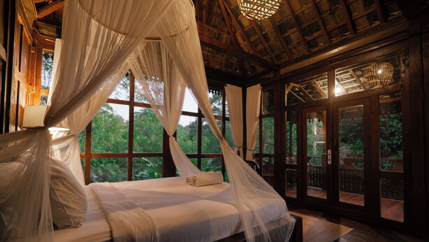 A bed with a canopy, large windows, and a view of greenery. - Kebun Kita Ecolodge (Pupuan)
