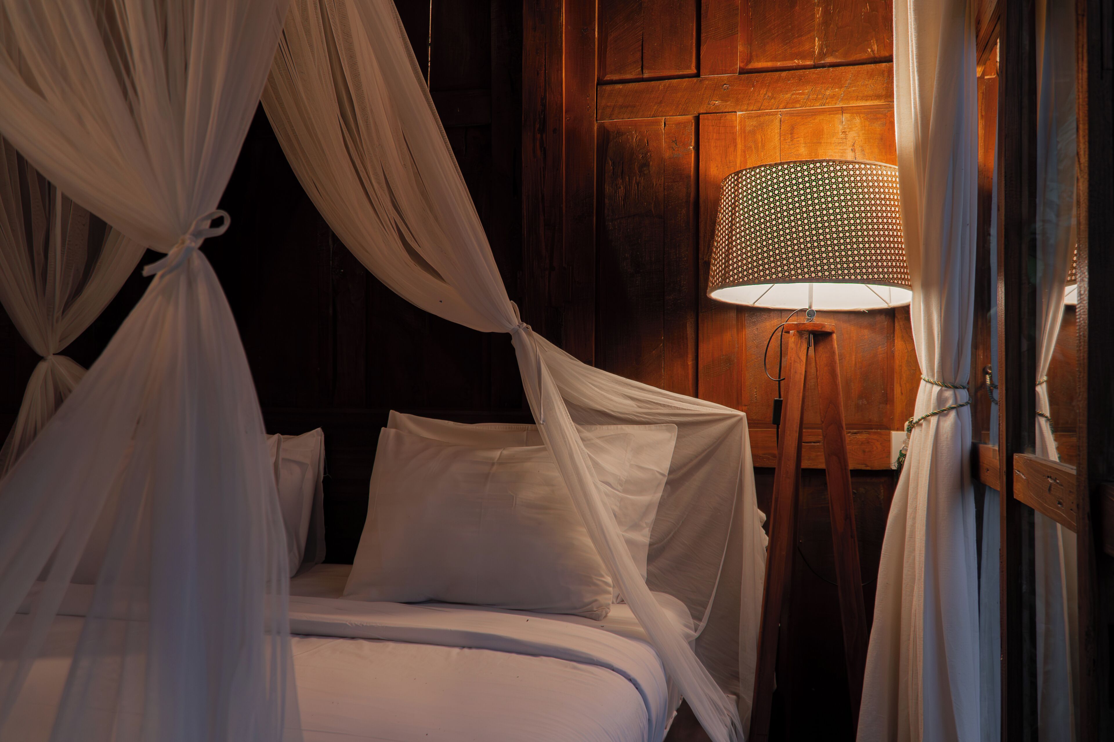 Una cama con ropa de cama blanca y dosel, una lámpara de pared y paneles de madera.
