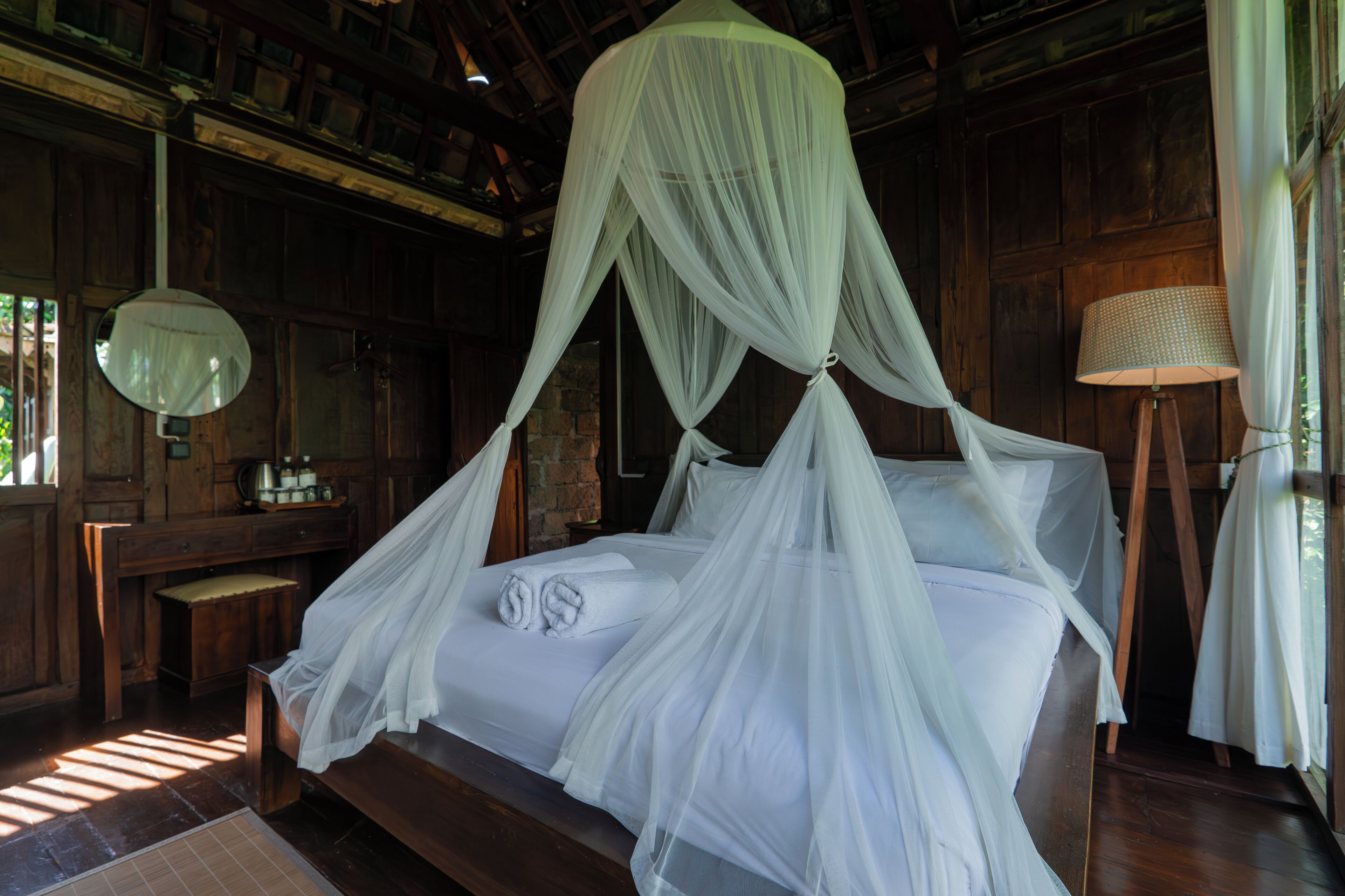 Una habitación de madera con una cama cubierta por un dosel blanco. Hay un banco de madera y una lámpara sobre una mesita.