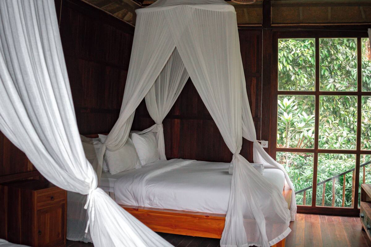 A room with a wooden ceiling and a large window. There is a bed with a canopy and a wooden bench.