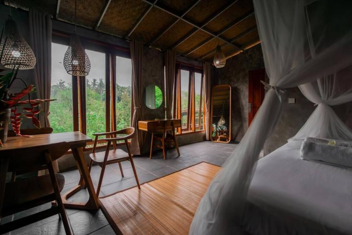 A room with a bed, a dining table, chairs, a mirror, and large windows with a view of greenery.