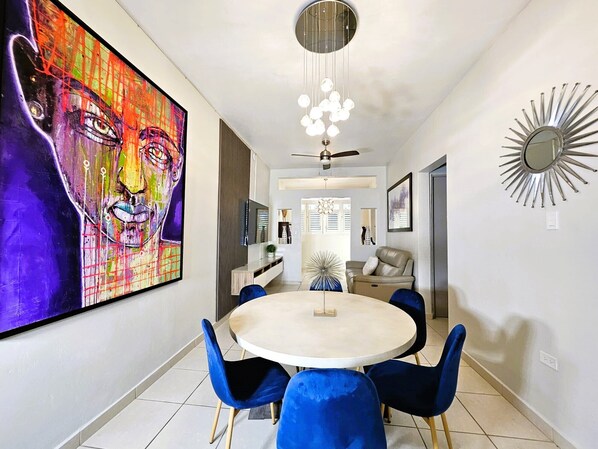 Uma sala de jantar moderna com uma mesa redonda, cadeiras azuis, um ventilador de teto e um grande quadro abstrato na parede.