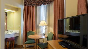 Habitación de hotel con televisor, una mesita redonda con sillas, lámpara, espejo y baño con lavamanos e inodoro.