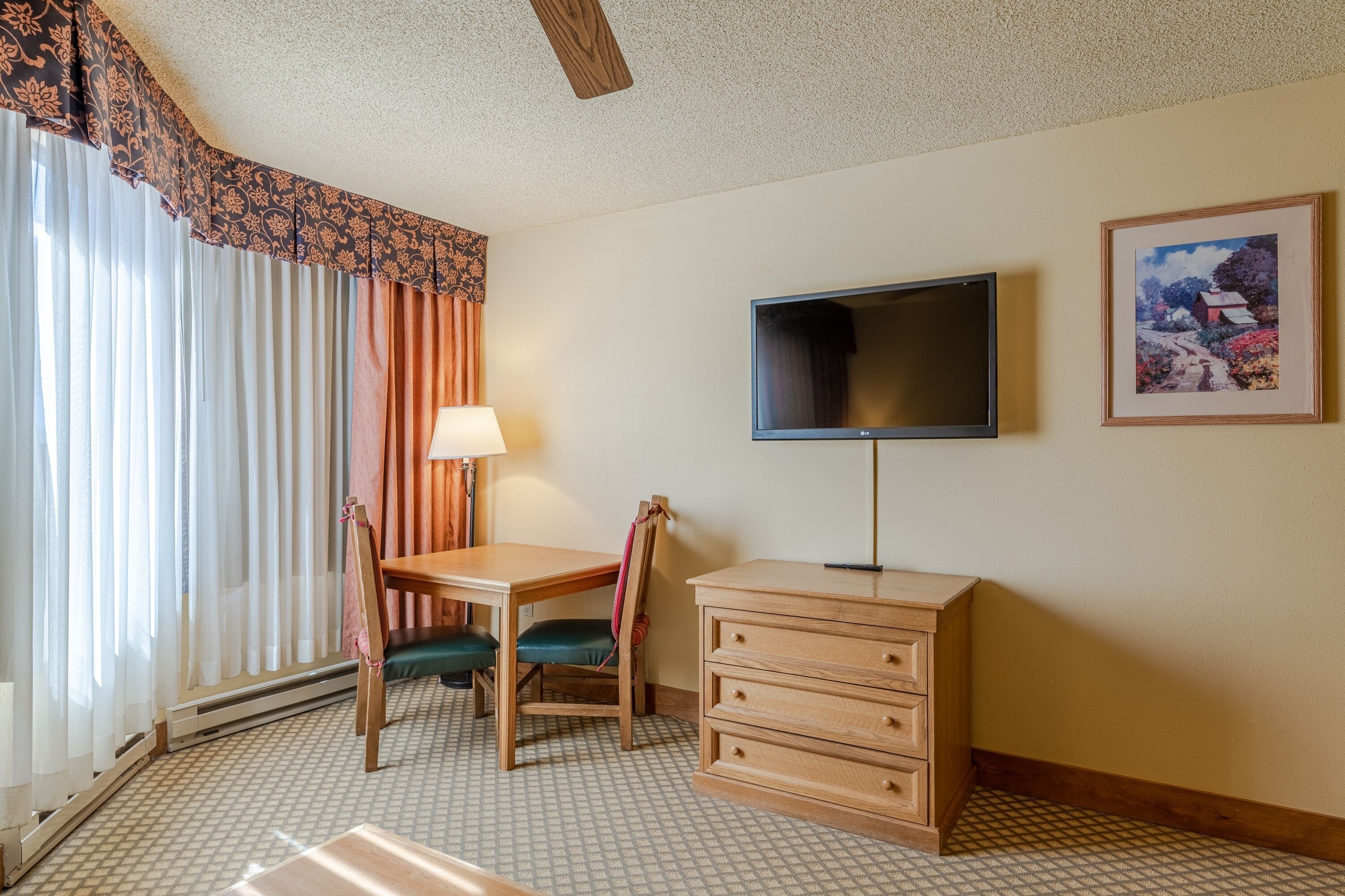 A hotel room with a bed, a desk with a chair, a dresser, a television on the wall, and a window with curtains.