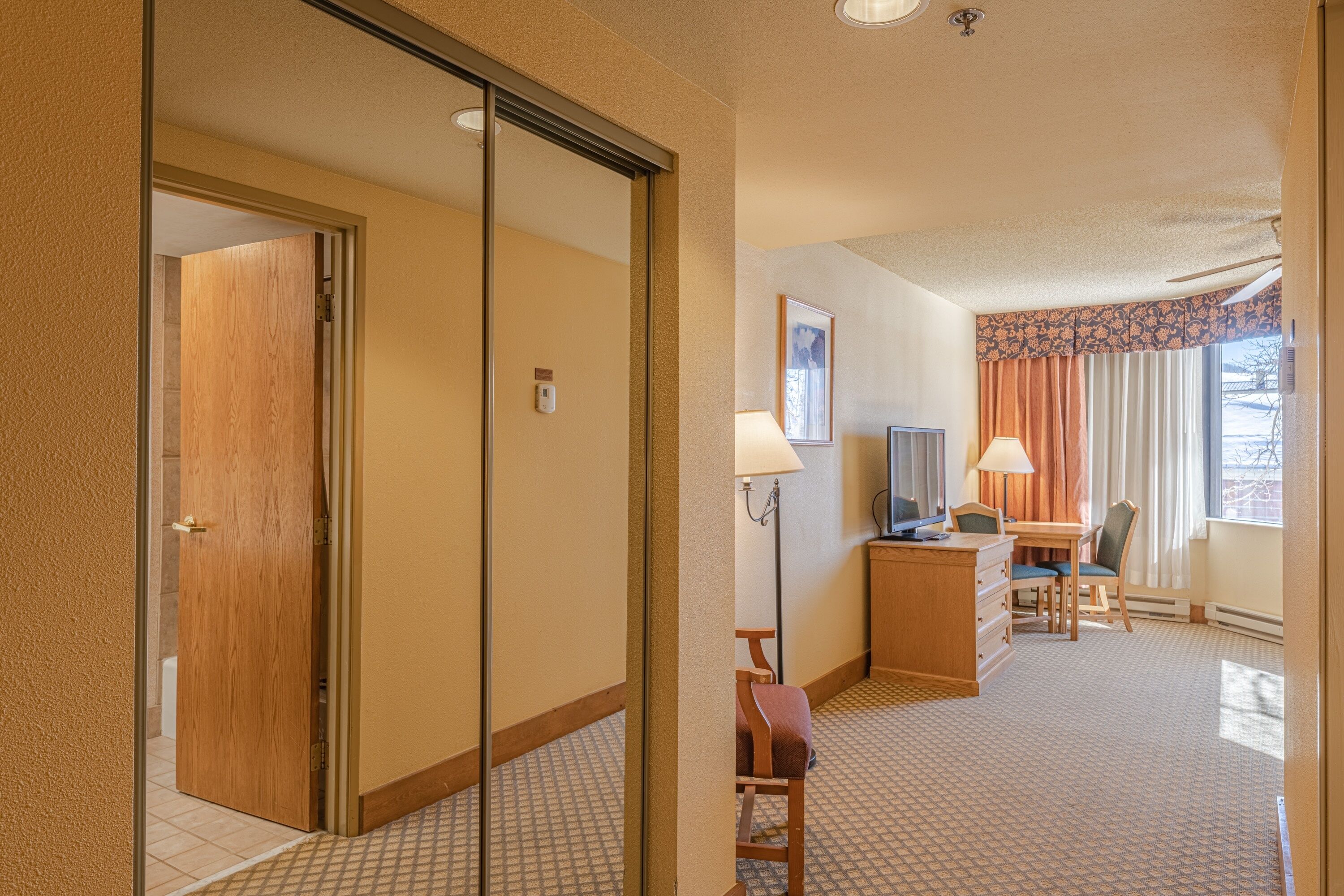 Ett hotellrum med en dörr av trä, ett skrivbord med en stol, en tv och ett fönster med gardiner.