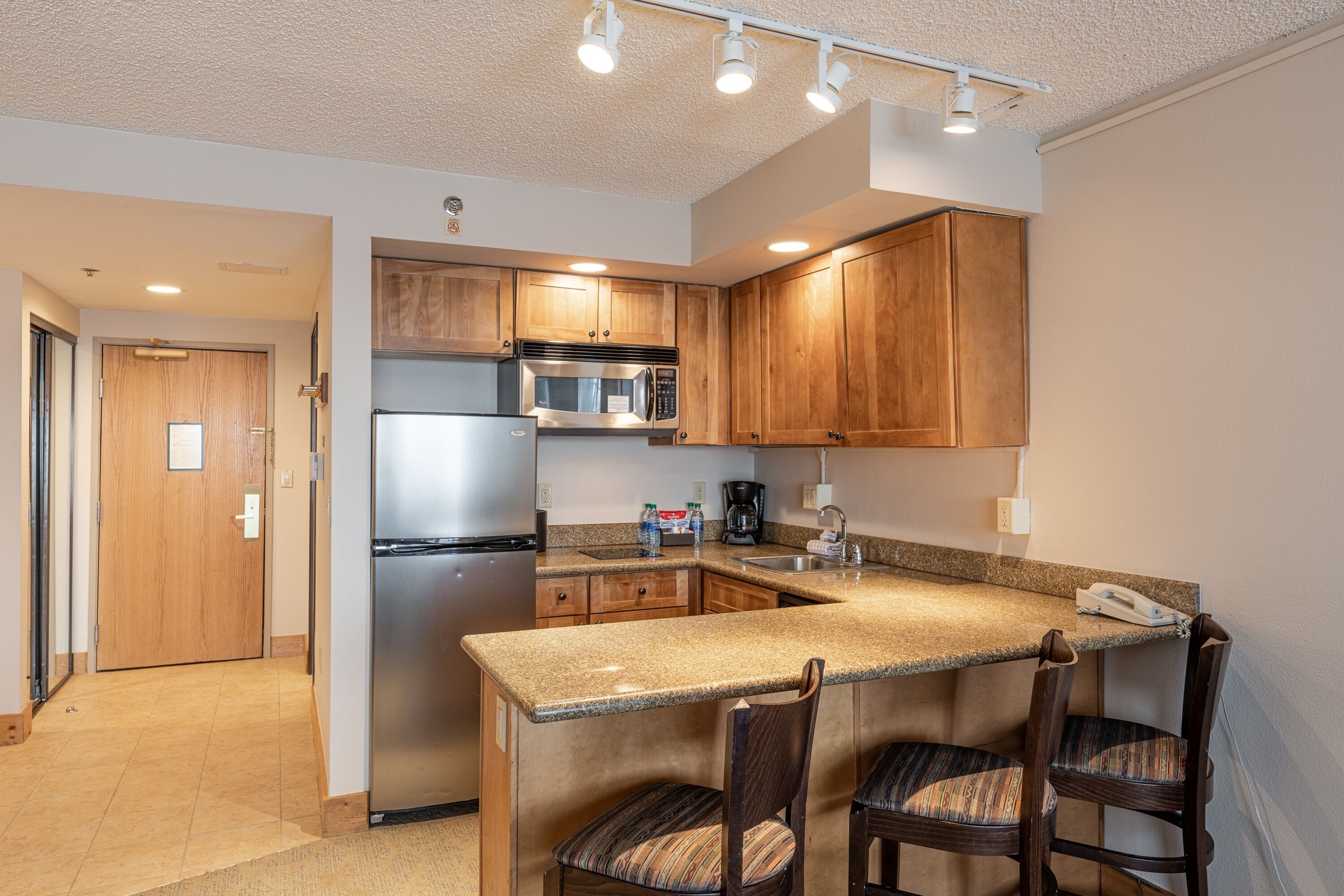 A modern kitchen with granite countertops, stainless steel appliances, wooden cabinets, and a bar area with two chairs.