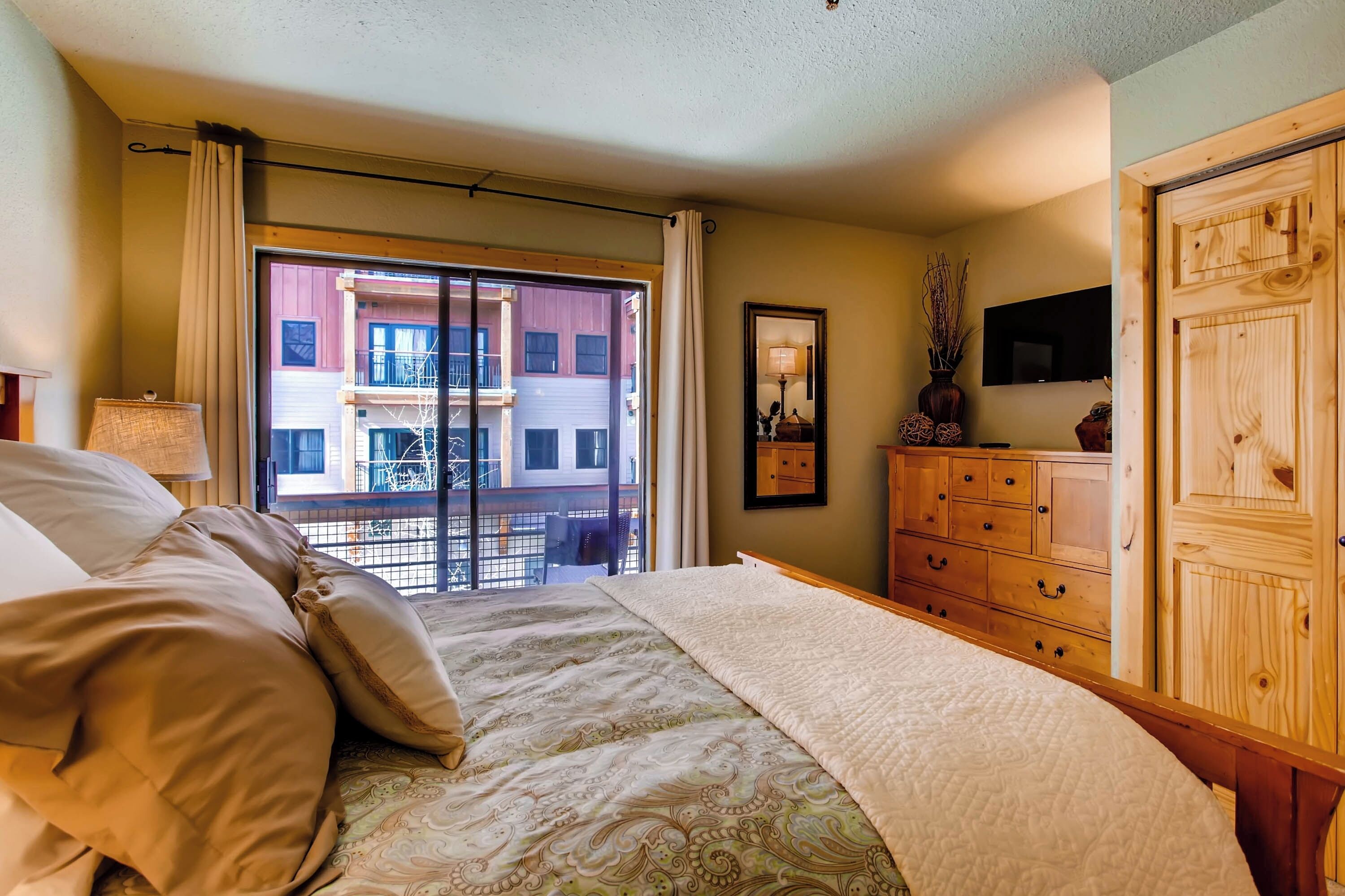 A bedroom with a bed, a dresser, a TV, and a view of a building outside.