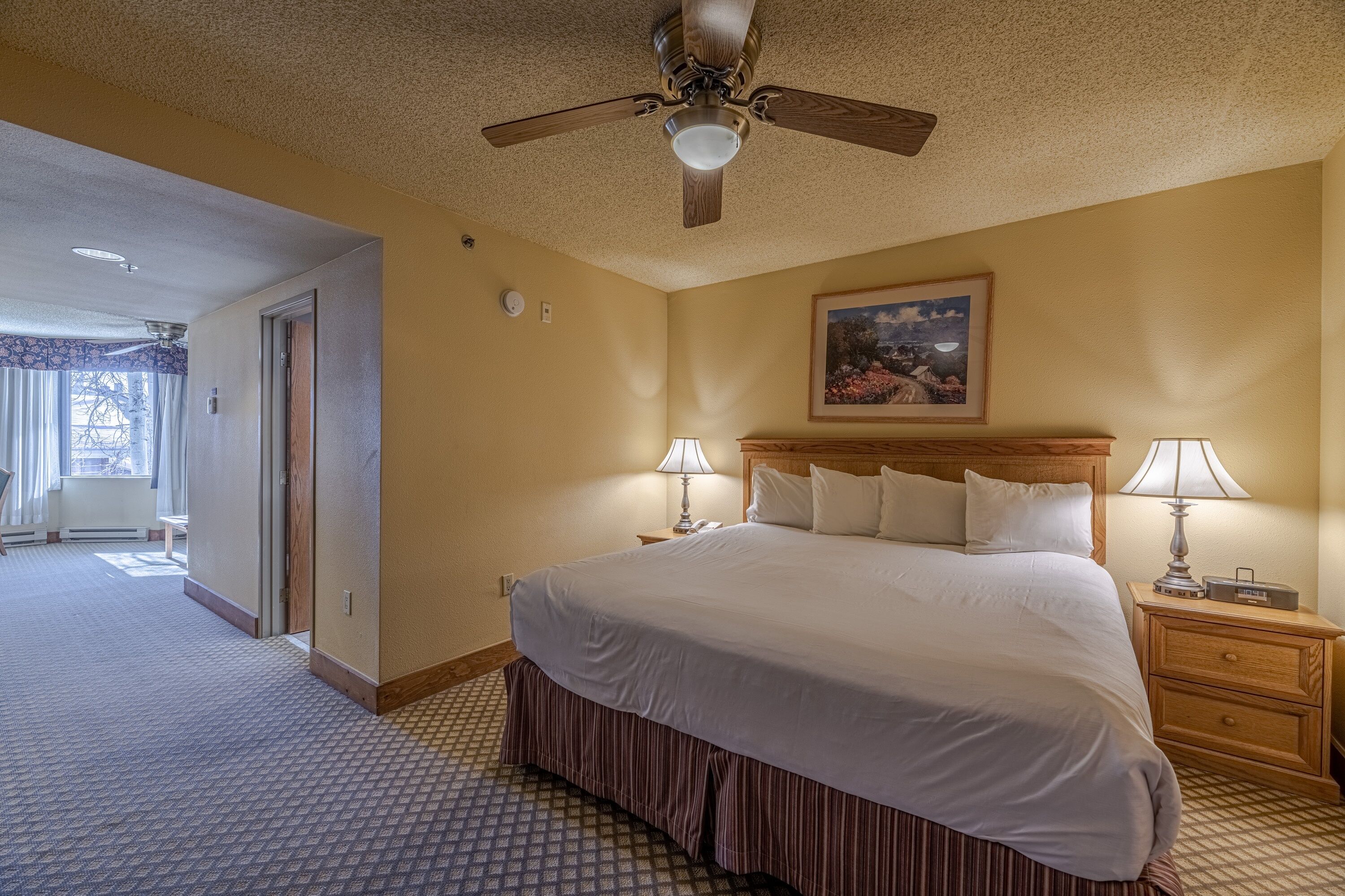 A hotel room with a large bed, two bedside tables with lamps, a ceiling fan, and a painting on the wall.