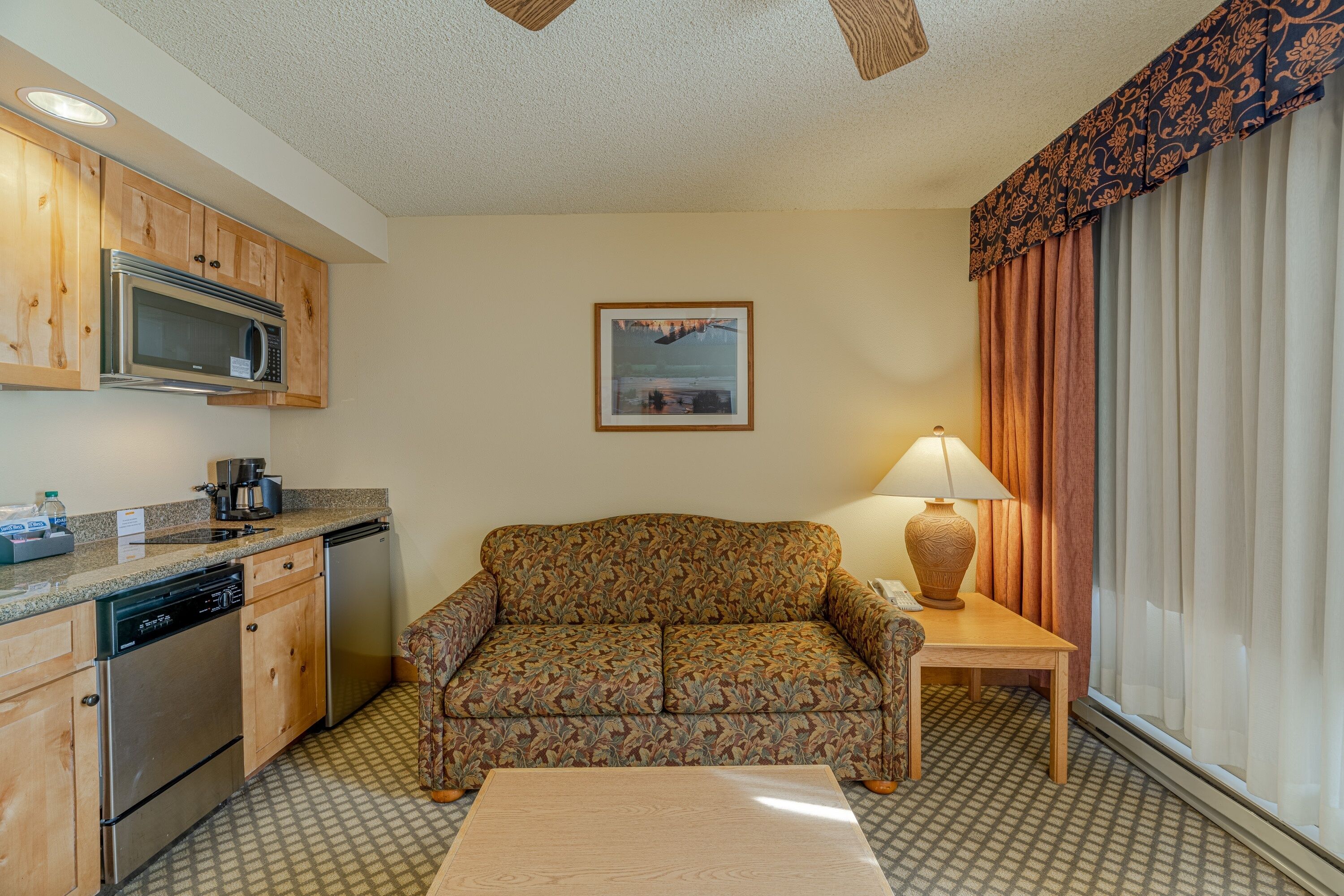 A compact hotel room with a kitchenette, a sofa, a table, and a window with curtains.