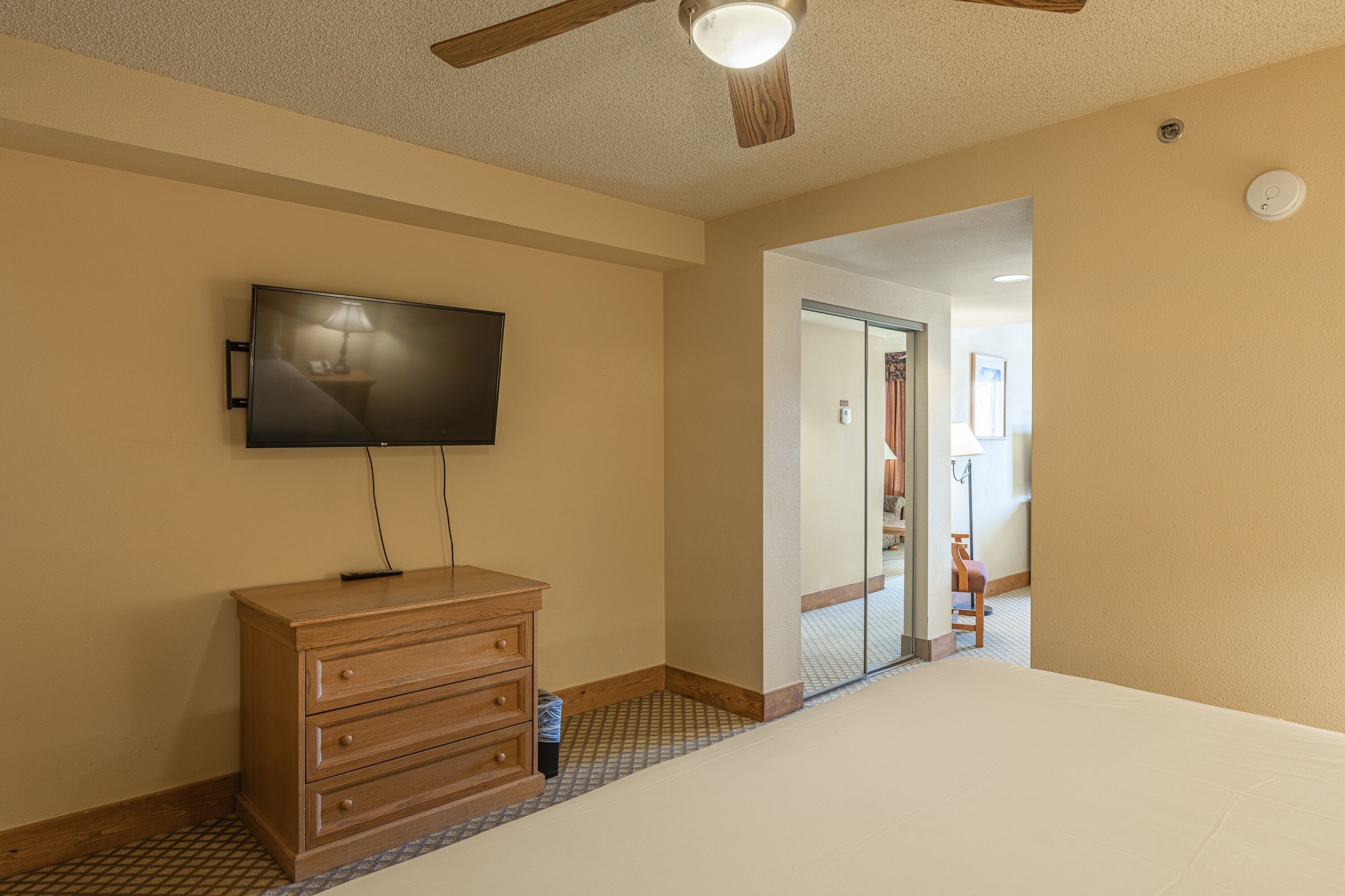 A hotel room with a flat-screen TV mounted on the wall, a wooden dresser, a ceiling fan, and a large bed.
