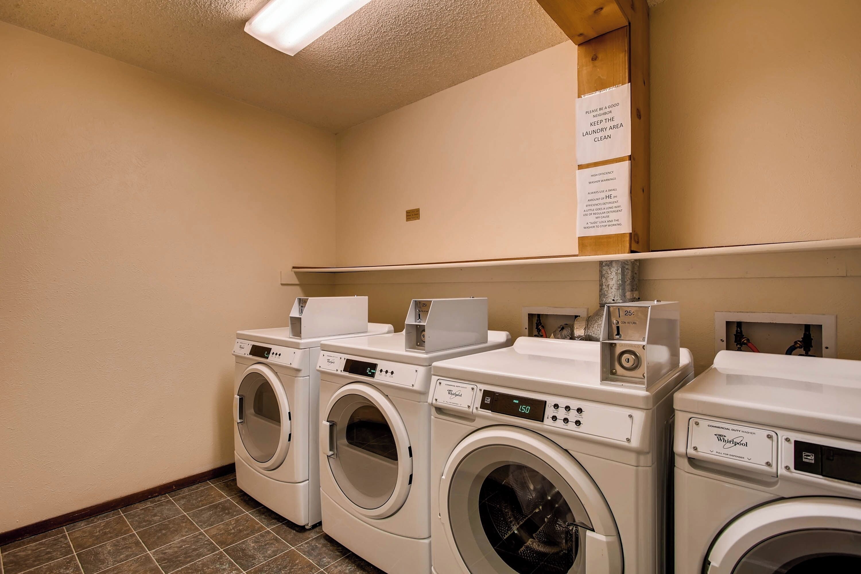 Une buanderie équipée de trois lave-linge et trois sèche-linge à chargement frontal, une étagère au-dessus avec une pancarte, et un sol carrelé.