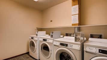 Une buanderie équipée de trois lave-linge et trois sèche-linge à chargement frontal, une étagère au-dessus avec une pancarte, et un sol carrelé.