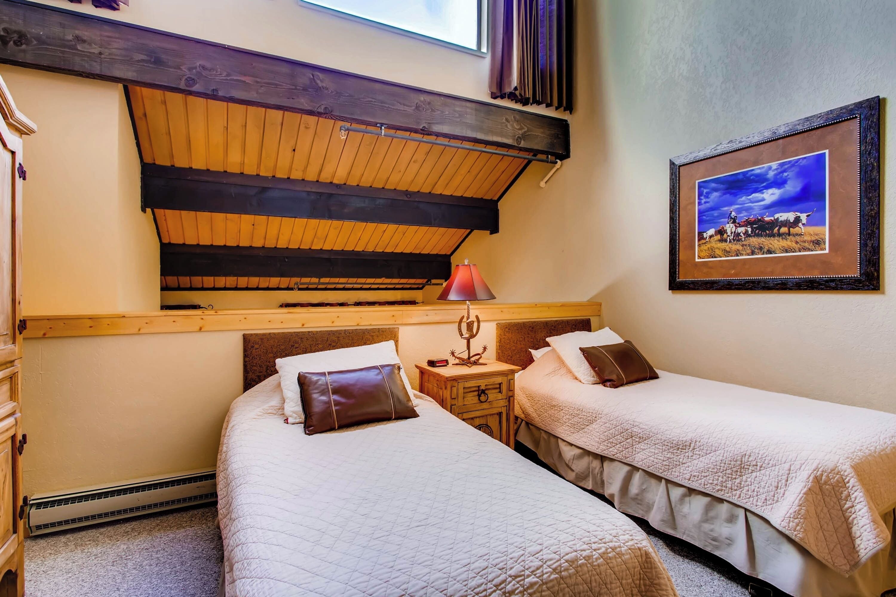 Una habitación con dos camas, techo de madera, un cuadro en la pared y una mesita con una lámpara.