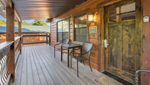 A wooden porch with a table and chairs, a wooden door with a welcome sign, and a view of a green-roofed building in the background. - Table Rock Lakeside cabin -Ticket Deals & Resort Perks included! (Branson)