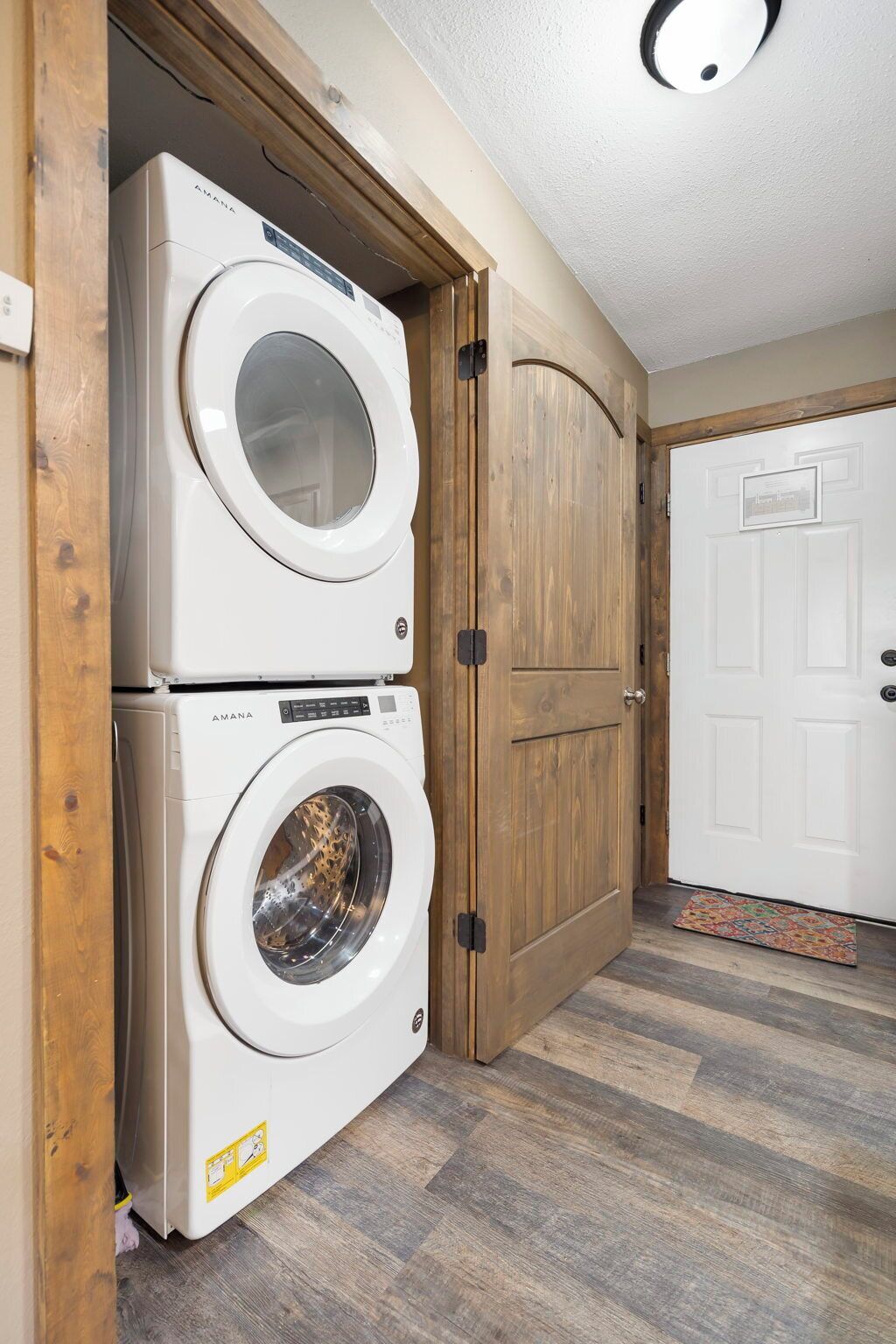 A laundry room with two stacked washing machines, wooden doors, and a patterned mat.