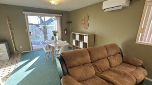Un salon avec un canapé marron, une table à manger avec des chaises, un petit meuble blanc et une fenêtre avec des stores.