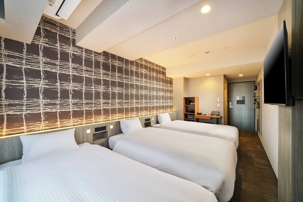 Ein Hotelzimmer mit drei Betten, einem großen, an der Wand montierten Fernseher und einem Schreibtisch mit Stuhl.