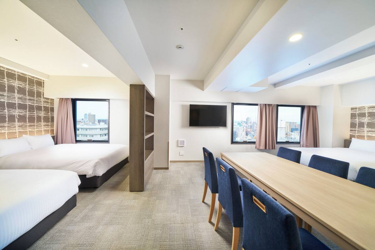 Ein Hotelzimmer mit zwei Betten, einem Esstisch, Stühlen, einem Fernseher und Blick auf die Stadt.