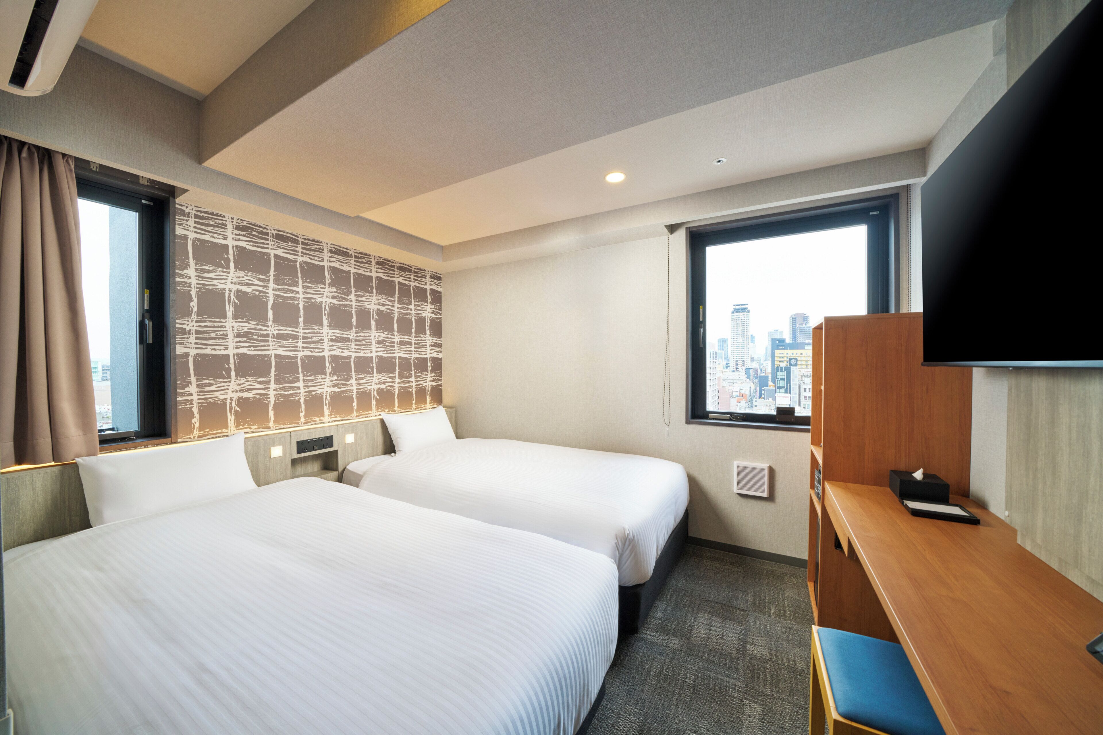 A hotel room with two beds, a desk, a TV, and a city view.
