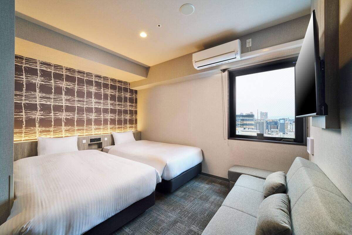Ein Hotelzimmer mit zwei Betten, einer Couch, Blick auf die Stadt und einer Klimaanlage.
