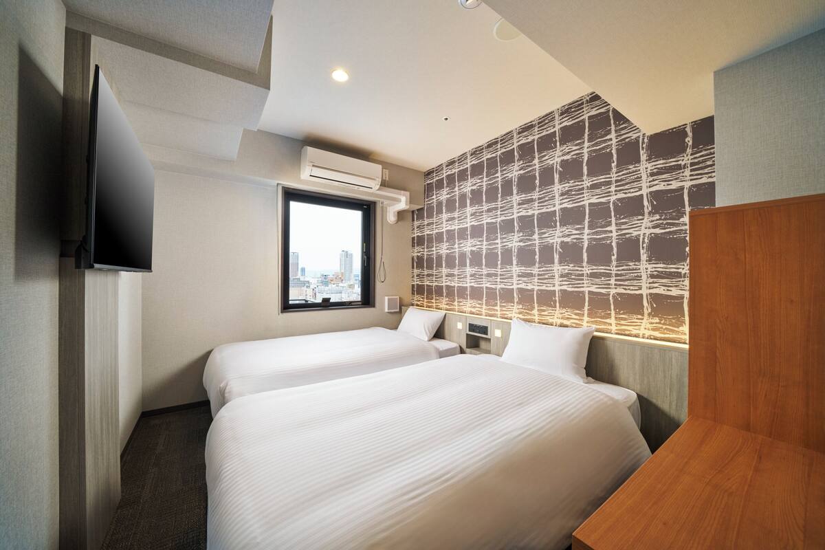 Ein Hotelzimmer mit zwei Betten, einem Flachbildfernseher, einer Klimaanlage und Blick auf die Stadt durch ein Fenster.