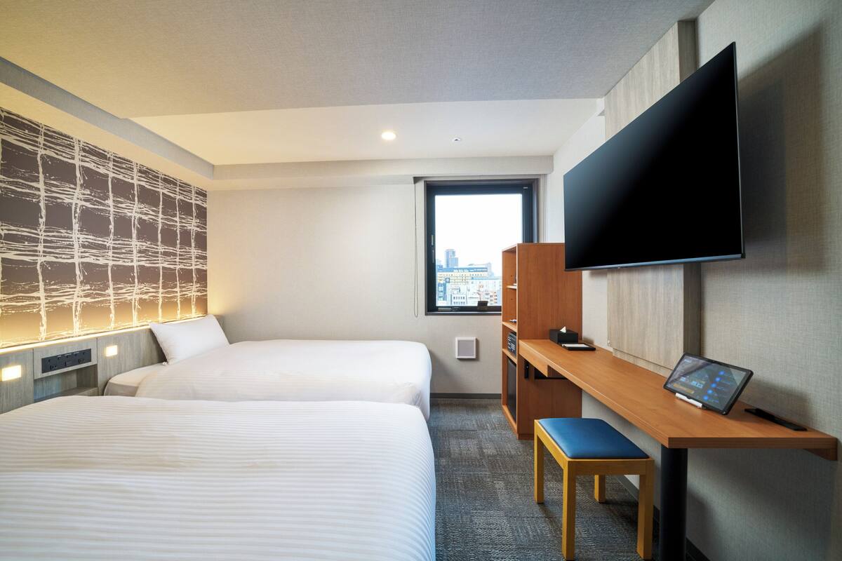 Ein Hotelzimmer mit zwei Betten, einem Schreibtisch mit Stuhl, einem Flachbildfernseher und einem Fenster mit Blick auf die Stadt.