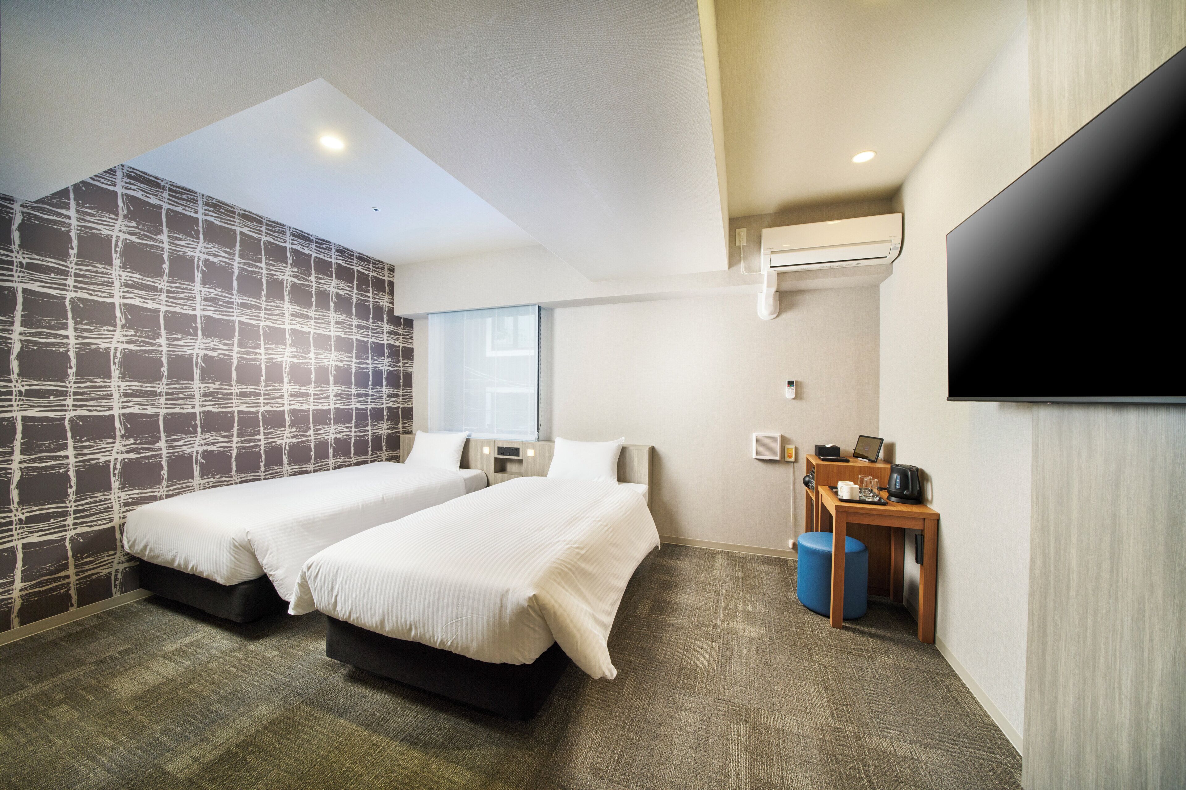A hotel room with two beds, a desk with a chair, a flat-screen TV, and a window.
