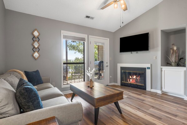 A living room with a sofa, coffee table, fireplace, TV, and a view through the glass doors. - Direct Lakefront Condo, sleeps up to 4, Resort Perks and Ticket Deals! (Branson)