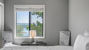 Un dormitorio con un ventanal grande, una mesita de noche con lámpara y almohadones decorativos.