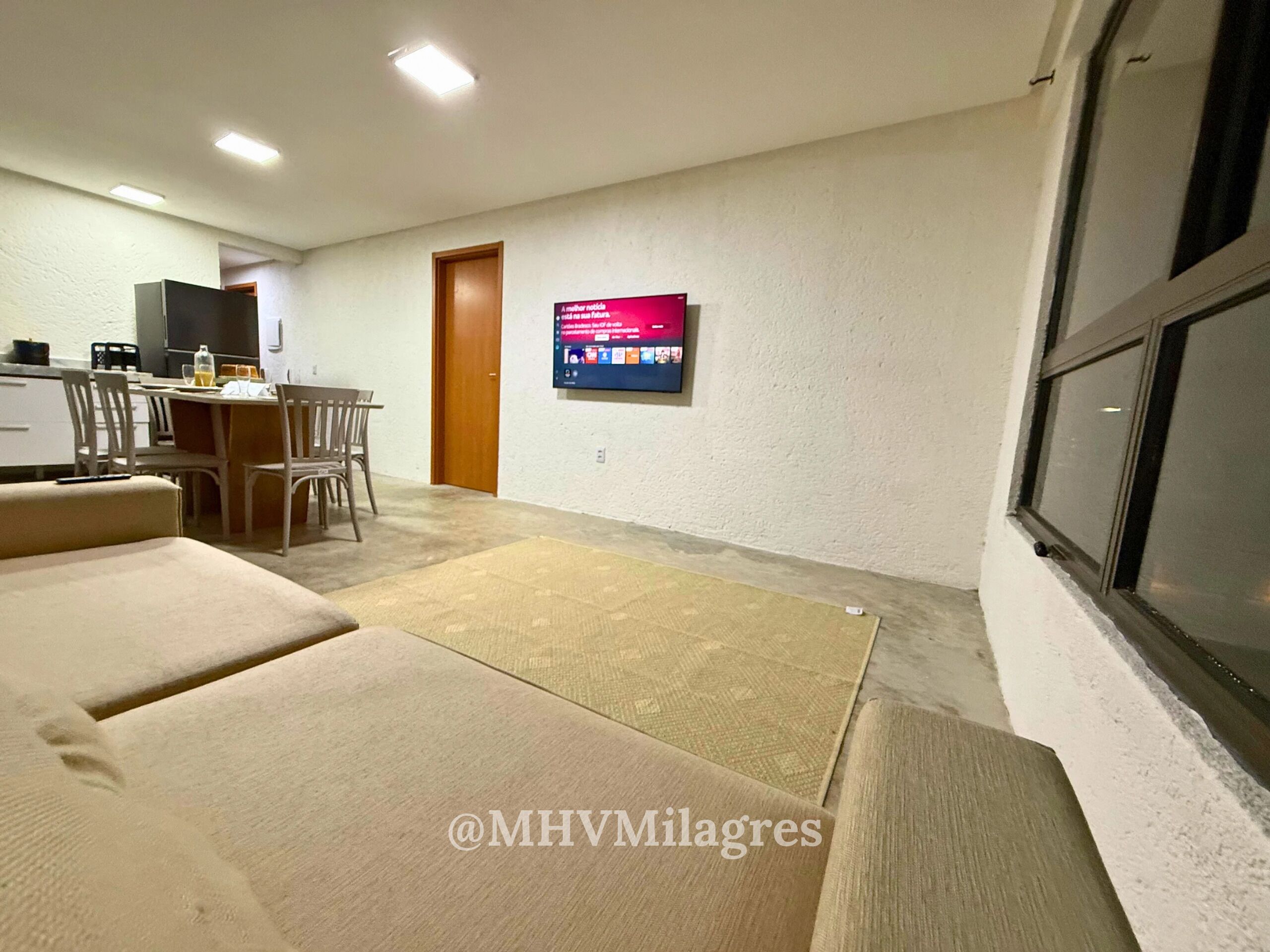 A living room with a sofa, a TV mounted on the wall, a dining area with chairs and a table, and a kitchen area with a refrigerator and a TV.