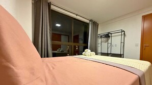 A hotel room with a bed, a window with curtains, a bedside table with towels, and a wall-mounted rack.