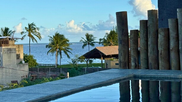 A pool with a view of the sea, palm trees, and a building with a tiled roof.