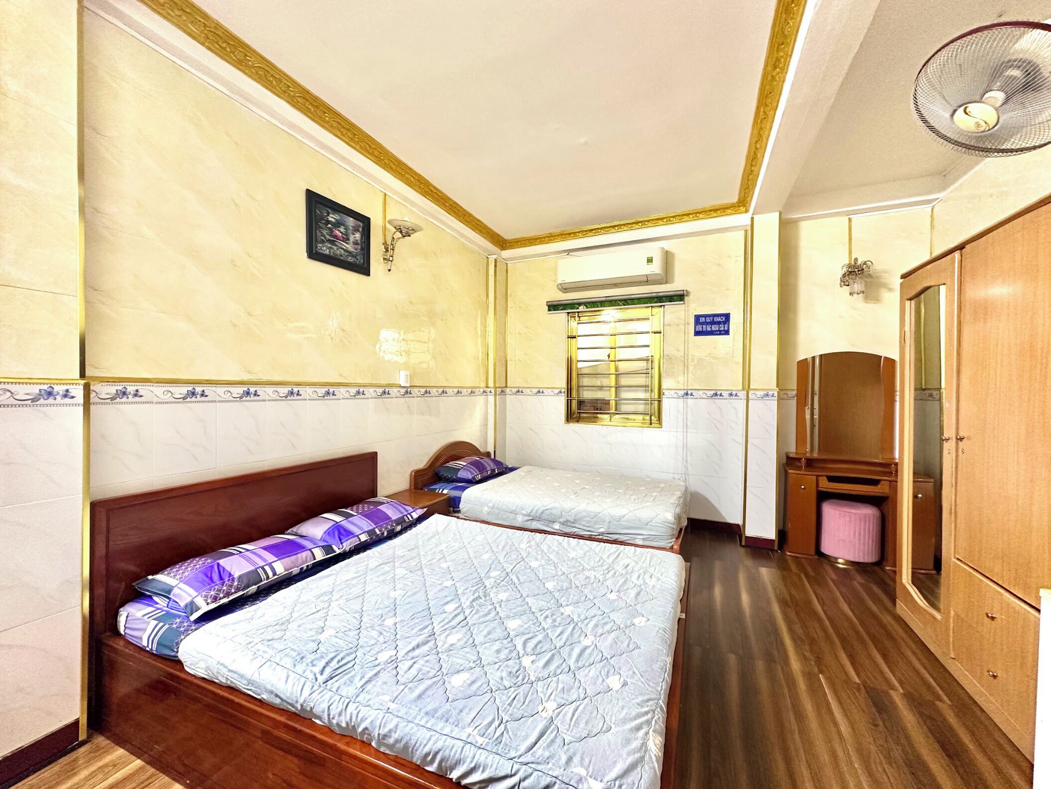 Hotellihuone, jossa on kaksi sänkyä, puinen vaatekaappi, peili, kattotuuletin ja ikkuna, jossa on sälekaihtimet.