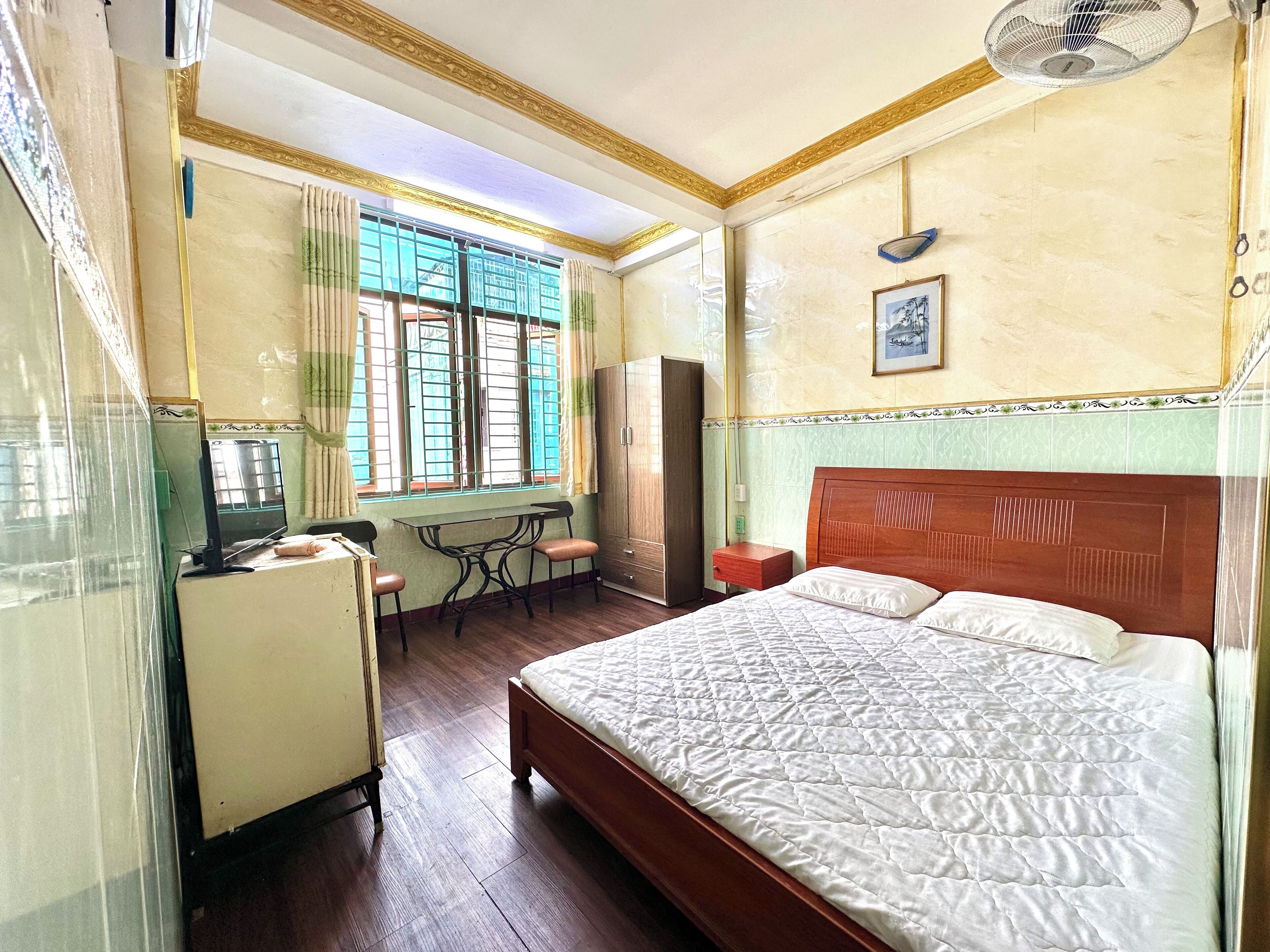 A hotel room with a bed, a desk with two chairs, a wardrobe, a window with curtains, and a ceiling fan.